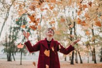 Stilvolle Frau in rotem Mantel wirft buntes Laub in Park und lacht — Stockfoto