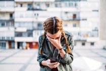 Руда дівчина з косами за допомогою мобільного телефону і обличчя з рукою проти житлового будинку — стокове фото
