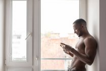 Smiling muscular black man using phone while standing against window in daylight — Stock Photo