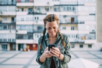 Smiling red-haired girl with braids using mobile phone against residential building — Stock Photo