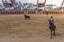Spanien, Tomelloso - 28. 08. 2018. Ansicht von Stierkämpfer, der Pferd reitet und mit Stier auf sandigem Gelände kämpft, mit Menschen auf der Tribüne — Stockfoto