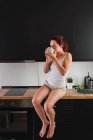 Frau sitzt auf Küchentisch und trinkt Kaffee in Küche — Stockfoto