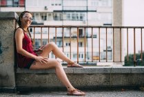 Souriant jeune femme rousse avec des tresses et des lunettes de soleil assis près de rambarde contre le bâtiment résidentiel — Photo de stock