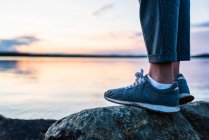 Crop side view of feet of person standing on stone near still lake on background of clear sky — Stock Photo