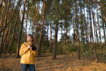 Mujer viajera contenido sosteniendo la cámara de fotos mientras está de pie en bosques de coníferas soleadas mirando hacia otro lado - foto de stock