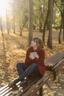 Attraente giovane signora in tenuta cappotto rosso aperto libro e seduto sul sedile nella foresta autunnale — Foto stock