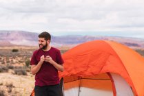 Бородатый парень в повседневной одежде держа кружку горячего напитка и современный смартфон, сидя рядом с палаткой и глядя прочь в красивой природе — стоковое фото