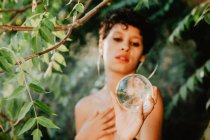 Jovem morena topless cobrindo peito e segurando bola transparente de vidro em madeiras verdes — Fotografia de Stock