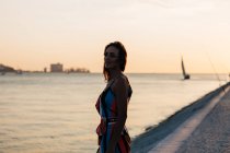 Verträumte junge Frau im Kleid steht bei Sonnenuntergang auf der Kopfsteinpflasterpromenade vor dem Meer und blickt auf die Aussicht — Stockfoto