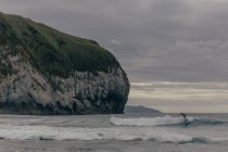 Persona surfeando en las olas altas en el mar agitado cerca de la roca enorme en el fondo del cielo gris sombrío - foto de stock