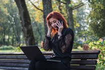 Жінка використовує ноутбук і смартфон на лавці в парку — стокове фото