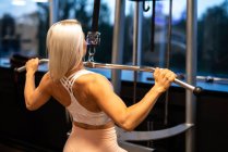 Bionda giovane donna in abbigliamento sportivo facendo Lat Tirare giù esercizio su attrezzature palestra vicino alla finestra — Foto stock