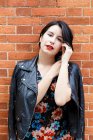 Jovem mulher bonita em jaqueta de couro e vestido com design de flor de pé perto da parede de tijolo e olhando para a câmera — Fotografia de Stock