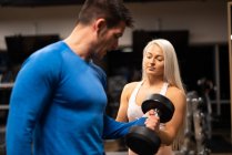 Mujer ayudando al hombre a hacer ejercicio con la mancuerna en el gimnasio - foto de stock