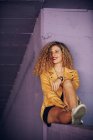 Trendy smiling curly woman in bright yellow jacket sitting on purple brick wall — Stock Photo