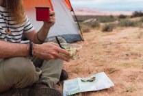 Erntehelfer essen Salat und genießen Heißgetränk, während sie auf sandigem Boden in der Nähe von Karte und Kompass beim Zelten in der Wüste sitzen — Stockfoto