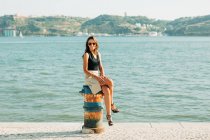 Trendy woman in high heels sitting on grungy stub on paved seafront — Stock Photo