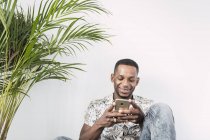 Sorrindo homem negro usando smartphone contra parede branca com planta verde — Fotografia de Stock