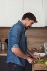 Vista lateral do jovem bonito cortando alface fresca para deliciosa salada enquanto está em pé na cozinha elegante — Fotografia de Stock