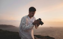 Handsome man holding photo camera and looking at pictures while standing on cliff in windy evening in San Francisco — Stock Photo