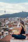 Боковой вид привлекательной блондинки в головном уборе, сидящей на крыше в позе медитации и смотрящей на живописный городской пейзаж — стоковое фото