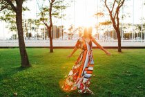 Cheerful stylish brunette in dress spinning around on green meadow in urban park — Stock Photo