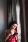 Молода жінка, спираючись на стіну біля вікна з мобільним телефоном — стокове фото
