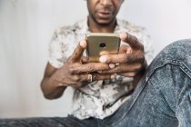Крупный план блестящего современного мобильного телефона в руках черного человека, сидящего у белой стены — стоковое фото