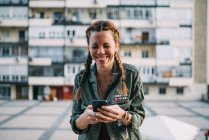 Сміється рудоволоса дівчина з косами, використовуючи мобільний телефон проти житлового будинку — стокове фото
