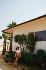 Mulher divertida vestida com camiseta branca e calções jeans com panamá segurando saco amarelo no fundo com casa de praia e palmas verdes — Fotografia de Stock
