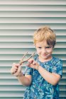 Blonde little boy in t-shirt playing with slingshot against shutters — Stock Photo