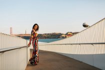 Улыбающаяся элегантная женщина в длинном платье, опирающаяся на забор моста в городе — стоковое фото