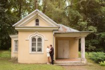 Vista lateral da noiva elegante e noivo abraçando perto de pequena casa velha no jardim verde — Fotografia de Stock