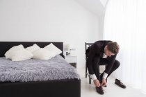 Junger Mann in schwarzem Kostüm und weißem Hemd sitzt auf Stuhl im Schlafzimmer und zieht Lederschuhe an — Stockfoto