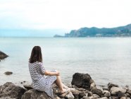 Mulher sonhadora sentado na costa rochosa e olhando para a vista — Fotografia de Stock