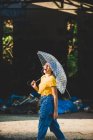 Encantadora mujer joven en traje elegante riendo y sosteniendo paraguas transparente mientras está de pie en la calle en el día soleado - foto de stock