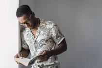 Casual black man in eyeglasses leaning on white wall near window and reading book — Stock Photo