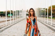 Contenu femme en robe ornementale colorée debout sur la promenade pavée et souriant à la caméra — Photo de stock