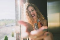 Young curly woman sitting in bright sunshine and outstretching blurred hand with smile — Stock Photo