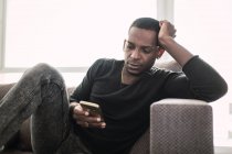 Випадковий афроамериканець людиною, сидячи на дивані у себе вдома і серфінгу смартфон спираючись з боку — стокове фото
