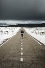 Back view of young person in stylish outfit walking in middle of asphalt road on cloudy winter day in beautiful countryside — Stock Photo