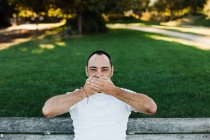Atraente adulto masculino cobrindo boca enquanto sentado no banco no parque no dia ensolarado — Fotografia de Stock