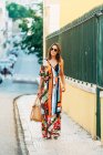 Mulher elegante em vestido longo carregando bolsa e andando na rua à luz do sol — Fotografia de Stock