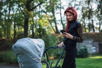 Young woman with ginger hair using smartphone while walking with baby stroller in park — Stock Photo