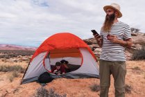 Maschio barbuto guardando lontano e navigando smartphone moderno mentre in piedi nel deserto vicino tenda e amico — Foto stock