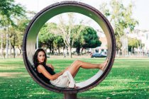 Ritratto di donna magra in gonna seduta in panchina rotonda contemporanea nel parco verde — Foto stock