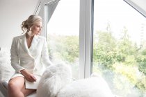 Young beautiful woman in white jacket with classic hairdo sitting on window sill with soft pillows and looking through window — Stock Photo
