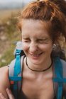 Mujer joven y bonita con la mochila sonriendo y entrecerrando los ojos mientras está de pie sobre un fondo borroso de la naturaleza en Bulgaria, Balcanes - foto de stock
