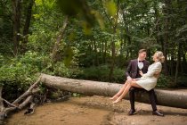 Handsome groom in suit holding bride on hands and sitting on fallen tree trunk in green woods — Stock Photo