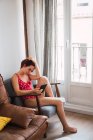 Jovem mulher de fato de banho vermelho sentado em poltrona no quarto e usando smartphone — Fotografia de Stock